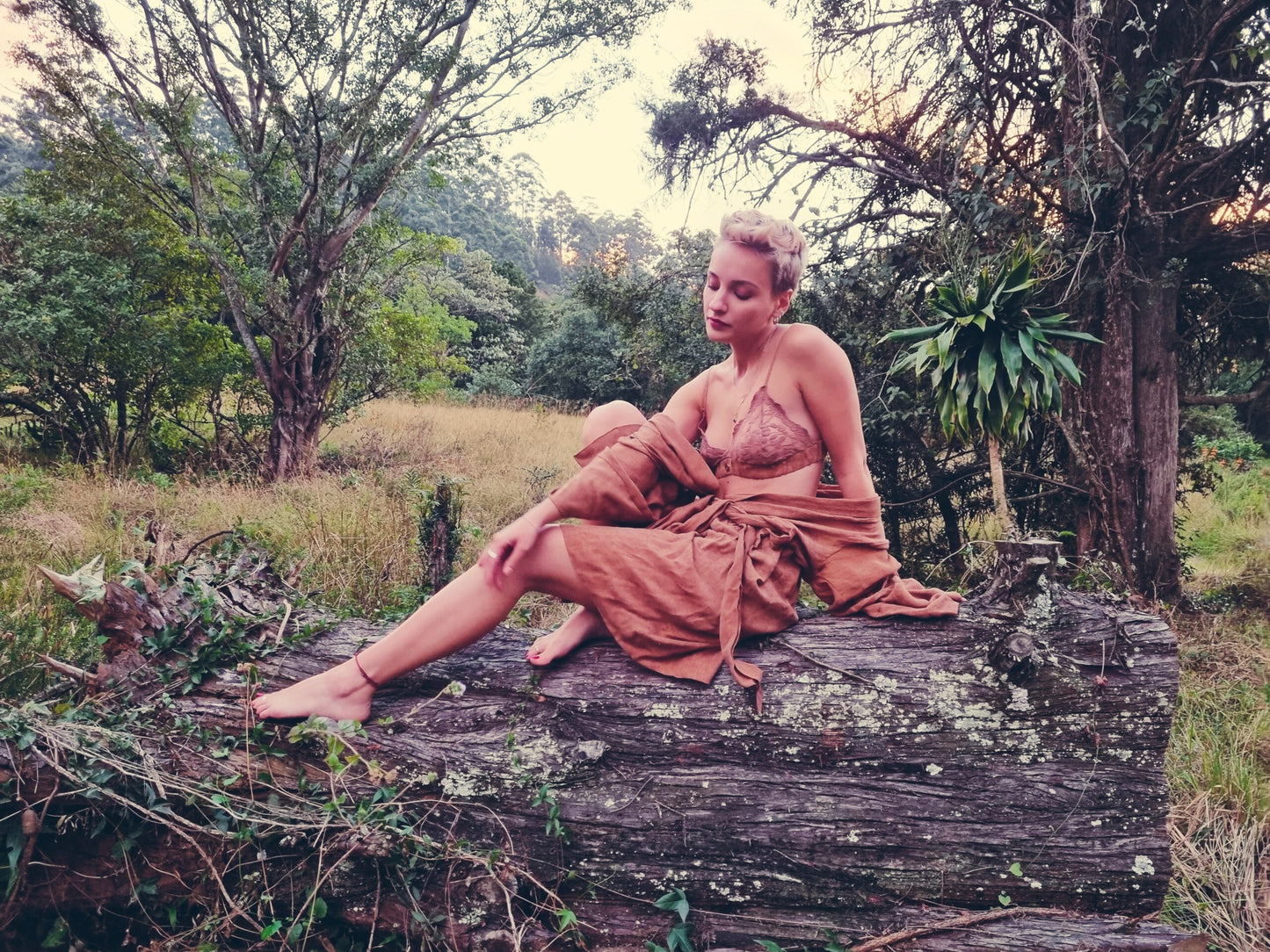 Short-haired stylish women sits outdoors on a log that has been cut down. She is wearing a rust coloured linen kimono and light pink lace bralet. The kimono is tied around her wait, but the shoulder have fallen down showing the bralet. As she sits she is touching her one leg while looking at the ground deep in thought. The items of clothing that she is wearing are from South African and are designed and made by hand in small quantities locally.