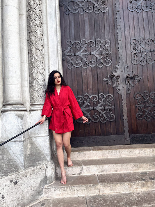  A sophisticated woman clad in a striking cherry red linen kimono descends the staircase of an elegant historical edifice. Barefoot and sporting alluring red lipstick complementing her attire crafted from premium linen fabric, her ensemble is meticulously handmade in South Africa.