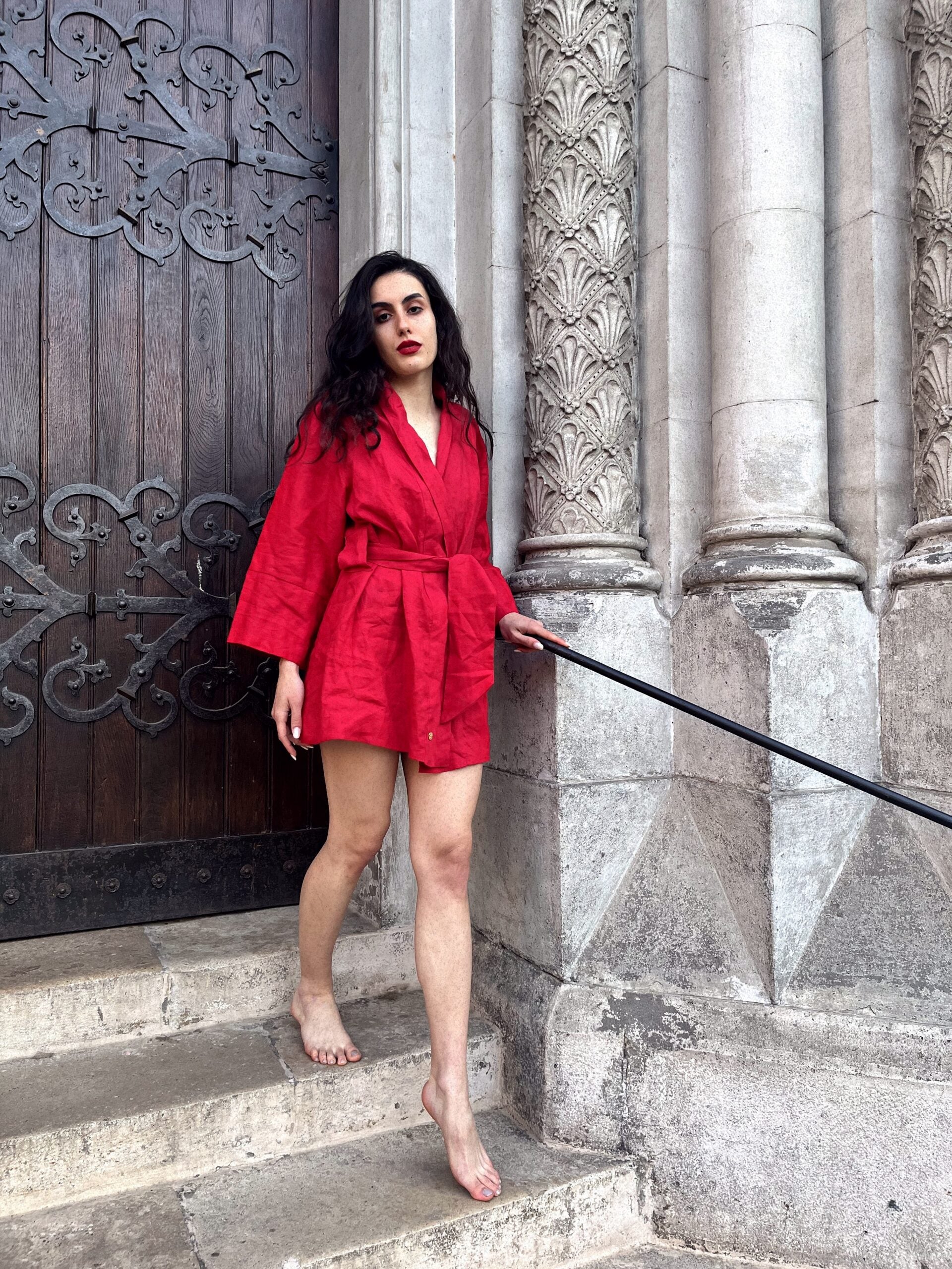 Classy lady wearing a vibrant chery red linen kimono walks down the steps of a grand old building. She is not wearing any shoes and is wearing seductive red lipstick to match her kimono. She is making eye contact with the camera. Her outfit is handmade from high quality linen fabric, and is designed and made in South Africa.