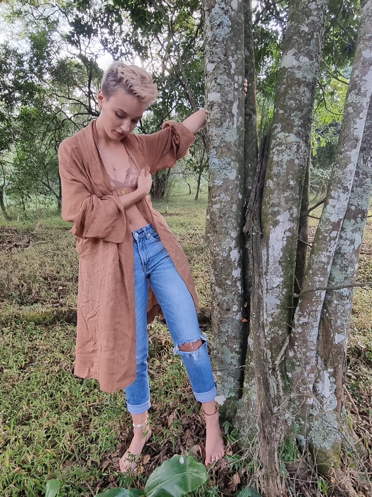 Stylish lady stands outdoors in nature alongside a tree wearing a rust coloured linen kimono, and light pink lace bralet and blue demin long pants. The linen kimono is hanging open showing the pink bralet that is worn underneath.