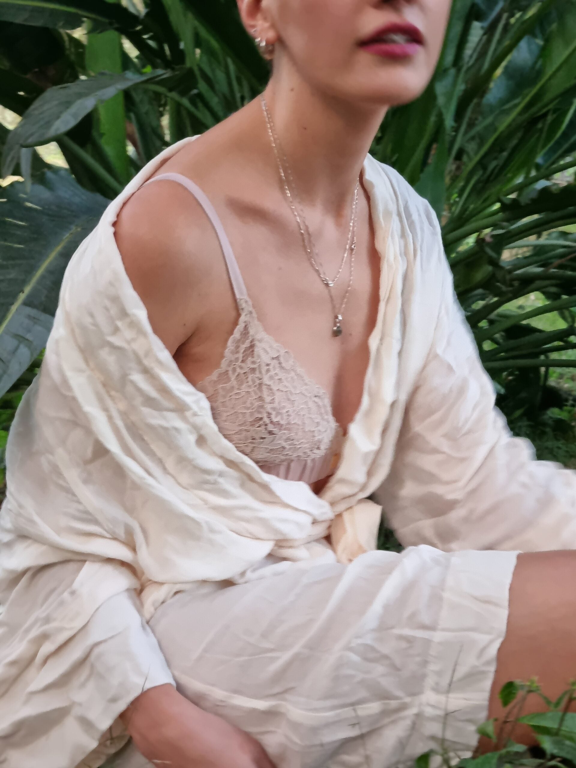 Woman sitting in her garden wearing a bralet and a white linen robe. The robe is falling off of her one shoulder, exposing the pink bralet on that side.