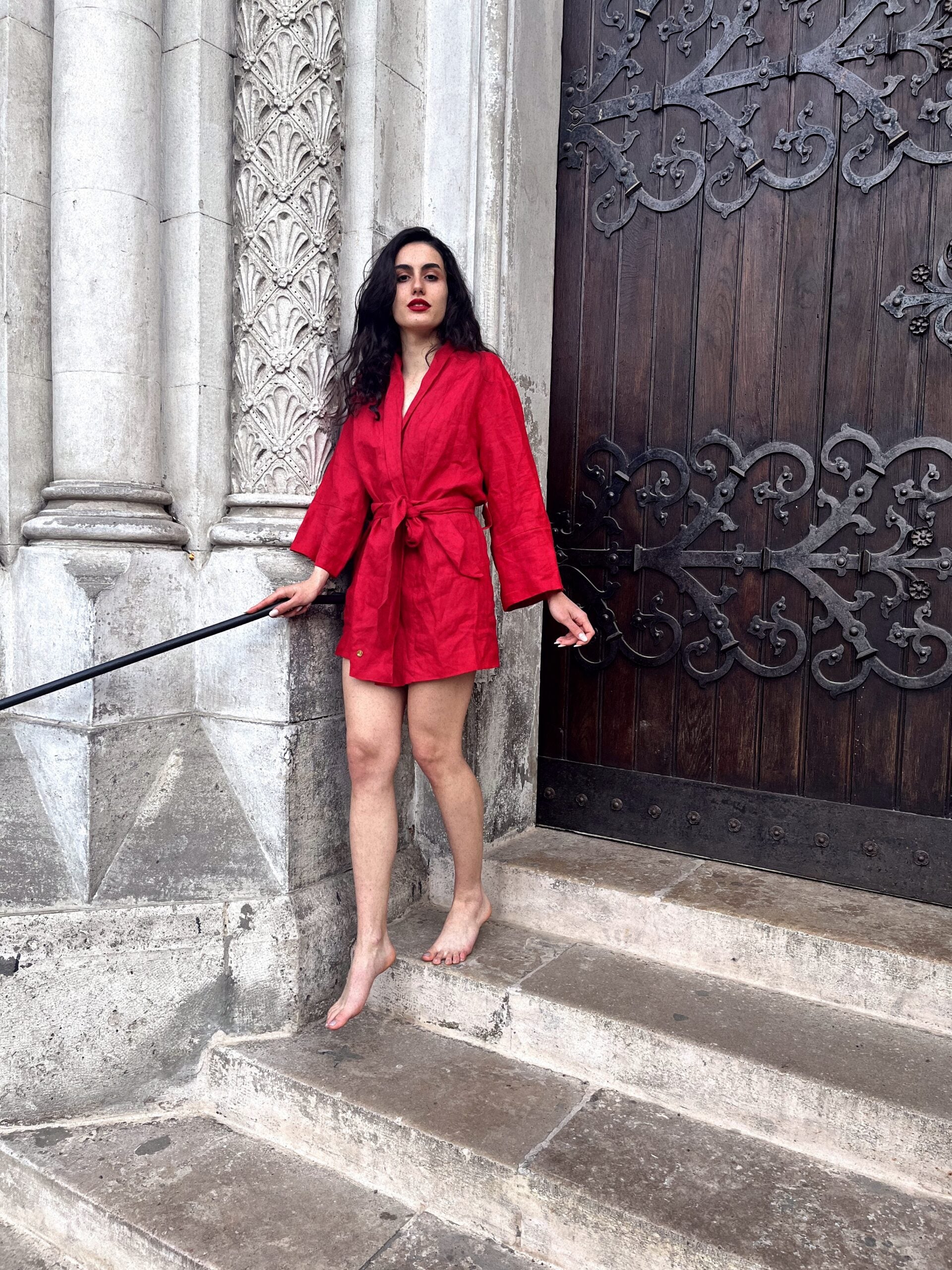 A stylish woman in a vibrant Chery red linen kimono gracefully stands on steps, leaning against a handrailing. Her attire is crafted by a renowned bespoke Linen clothing designer from South Africa.