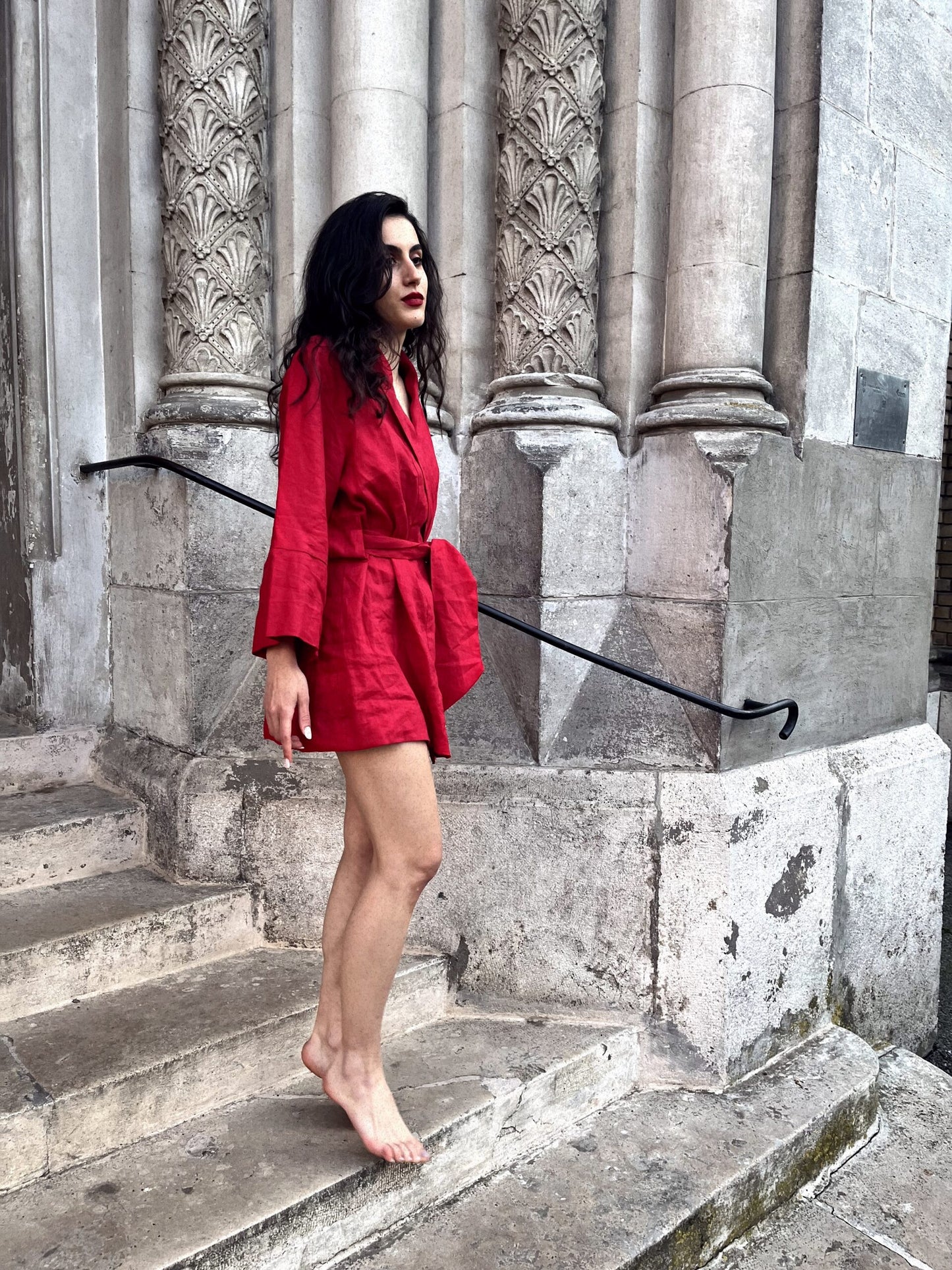 Sophisticated powerful woman walks down the steps of a glamorous old building while wearing a vibrant chery red linen kimono. She is barefoot with her legs seductively showing as she stares ahead into the distance. Her kimono is a bespoke handmade garment designed and manufactured in South Africa.