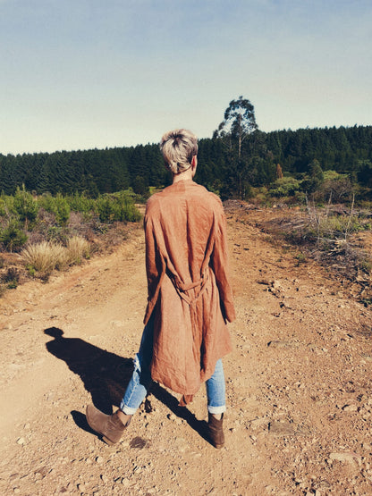 Stylish women wearing a rust coloured kimono, blue jeans and leather boots stands in the middle of a gravel road facing away from the camera. Her bespoke linen kimono is untied in the front and hangs down as she stands. The kimono she is wearing is made in very limited quantites with very high quality materials by South African linen clothing and loungewear experts Hare and Manuela