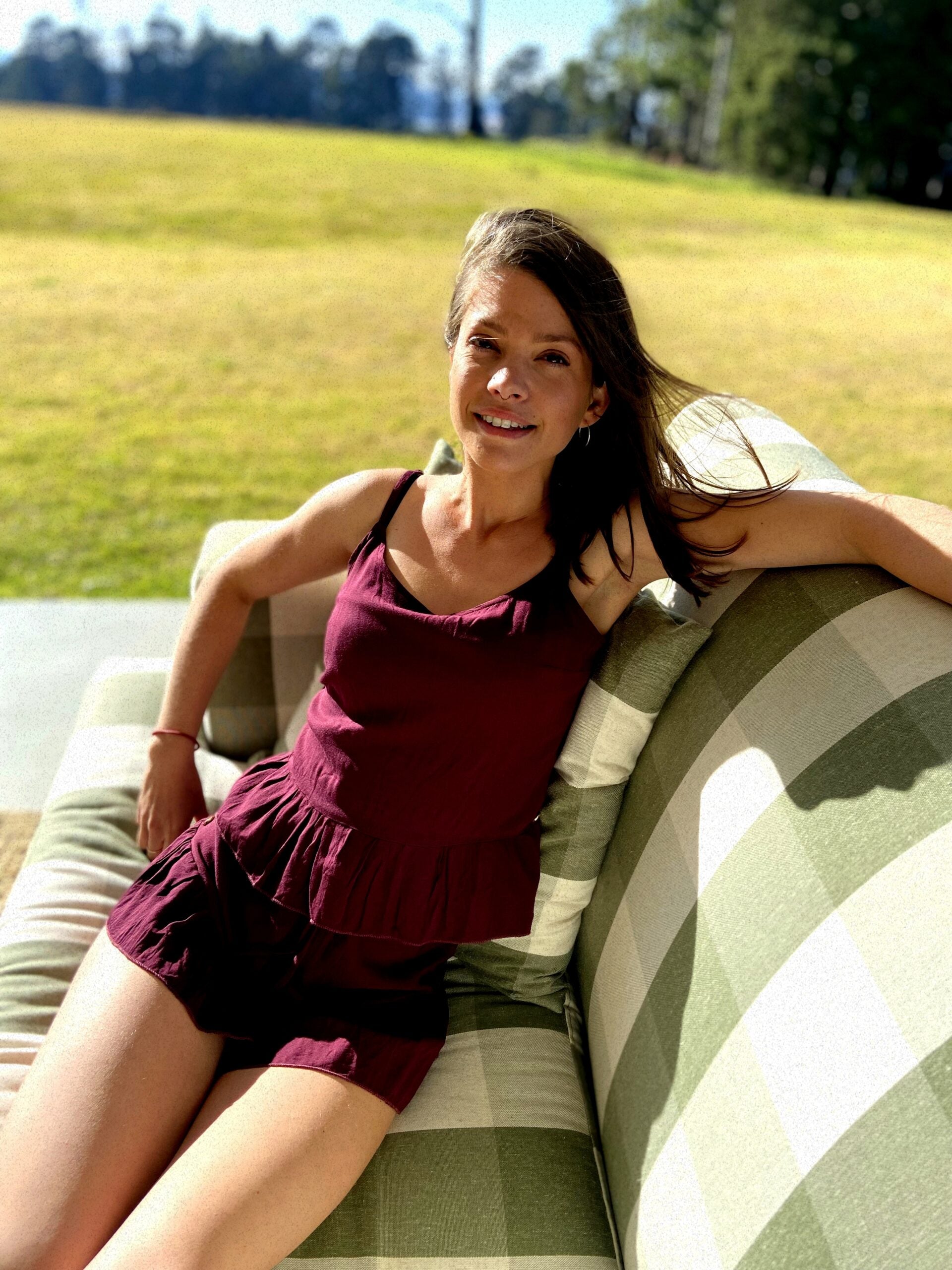 Woman lounges in the sun on an outdoor couch while wearing a bespoke handcrafted linen loungewear set made up of a plum coloured linen top and plum coloured linen shorts. 