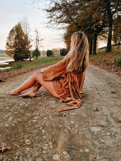 Women sits outdoors on a gravel road wearing a rust coloured kimono made of line overtop of her rust coloured linen pyjama set. The clothing items she is photographed wearing are manufactured by hand in limited quantities by South African linen clothing and loungewear designers. 