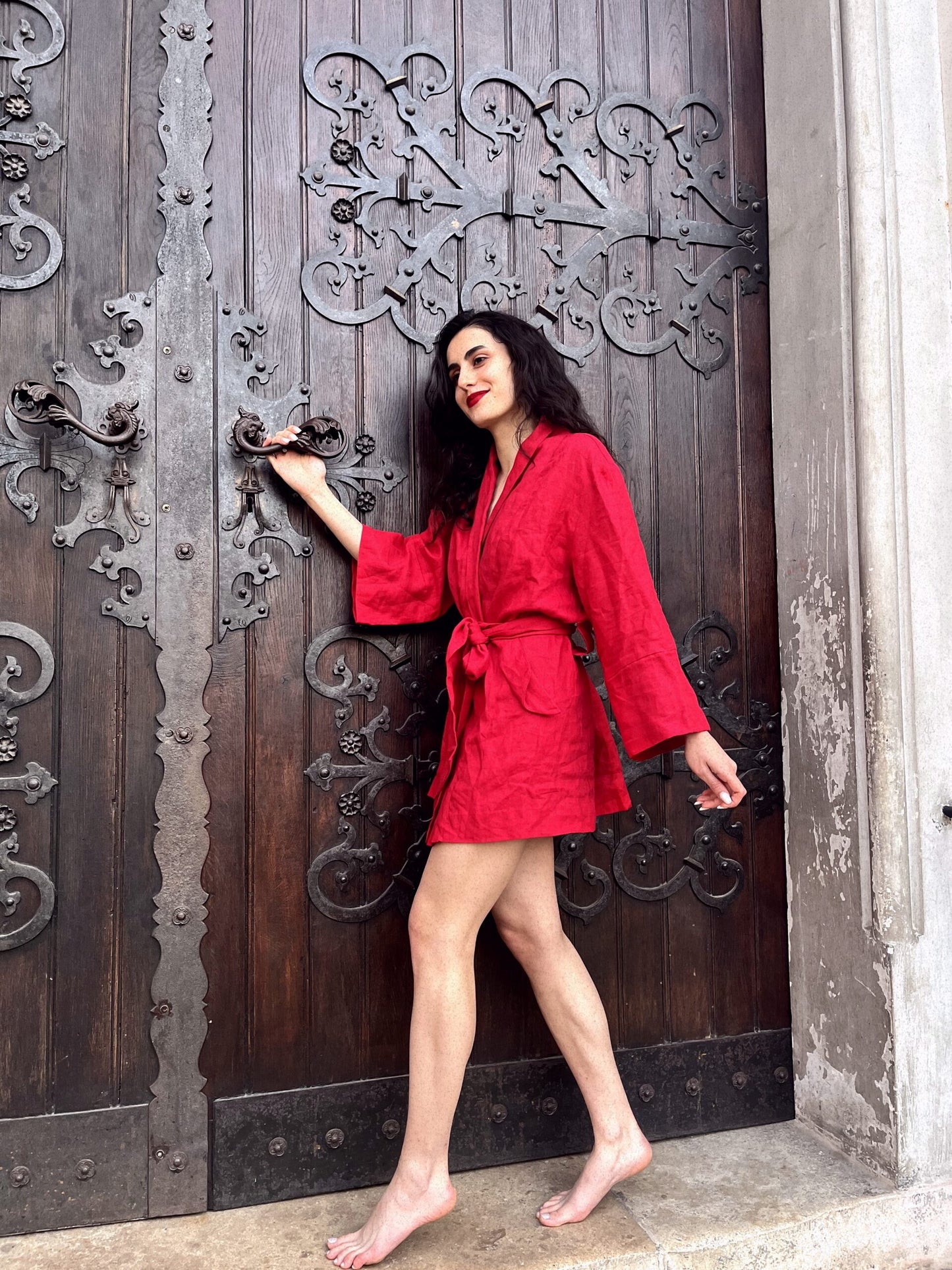 Adorned in a captivating Chery red kimono made of linen, a woman exudes elegance as she stands on steps holding onto a large old fashioned wooden door. Her exquisite clothing is meticulously crafted by a bespoke Linen clothing designer based in South Africa.