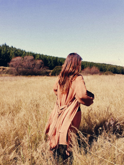Beautiful woman walks through a field of long grass wearing a rust coloured kimono made of linen. The woman is walking away from the camera, and her kimono is hanging open and flows as she walks. The kimono worn is made of linen and manufactured by hand in small limited quantities by South African linen clothing and loungewear manufacturers