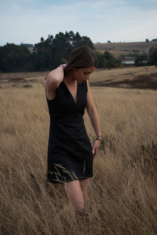 Woman stand outdoors in a grassy field wearing a little black dress made out of luxury linen fabric.