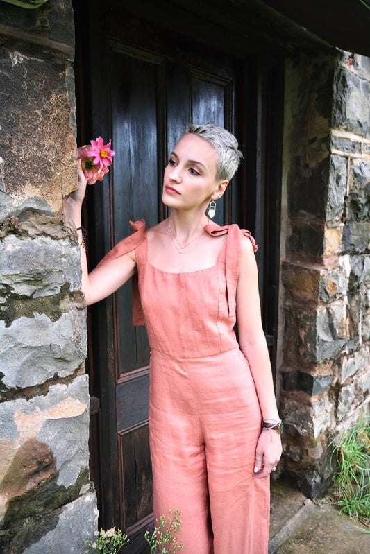 A stylish woman in a bespoke pink jumpsuit exudes elegance as she leans against a rustic stone wall, showcasing the craftsmanship of South African linen clothing.