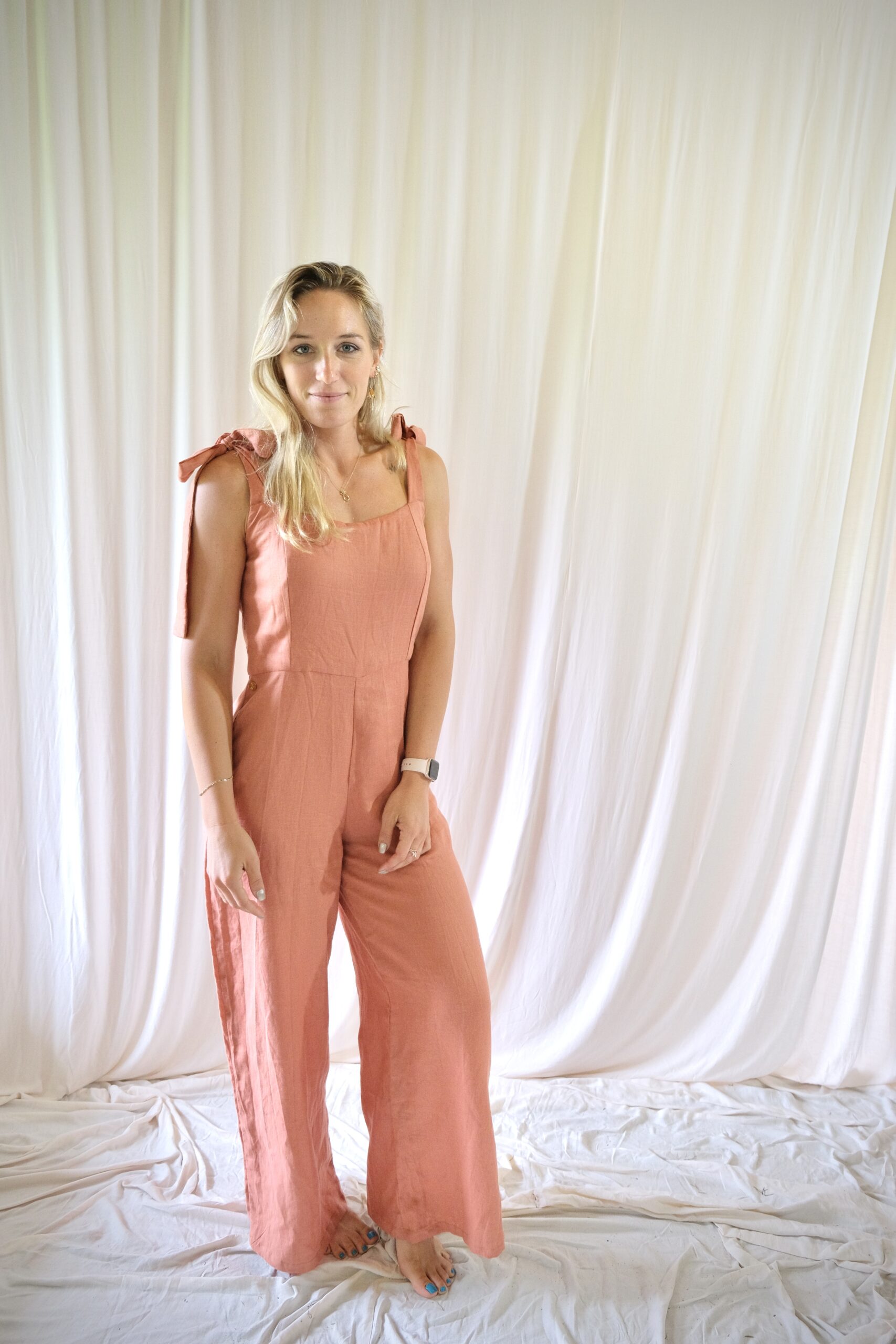 Embodying sophistication, a woman clad in a custom-made pink jumpsuit stands powerfully in front of a flowing white backdrop, showcasing the impeccable craftsmanship of South Africa's renowned linen clothing designer.