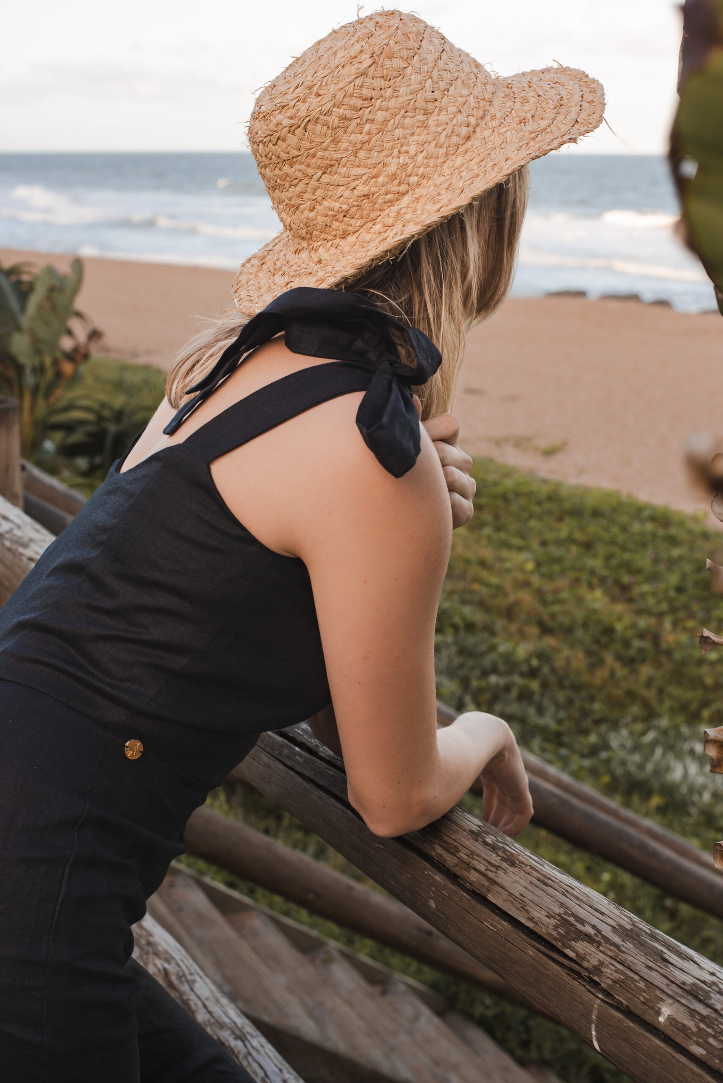 Woman wearing a bespoke linen black jumpsuit leans on boardwalk railing alongside the beach. She is wearing a straw hat and black jumpsuit and staring out into the distance deep in thought.
