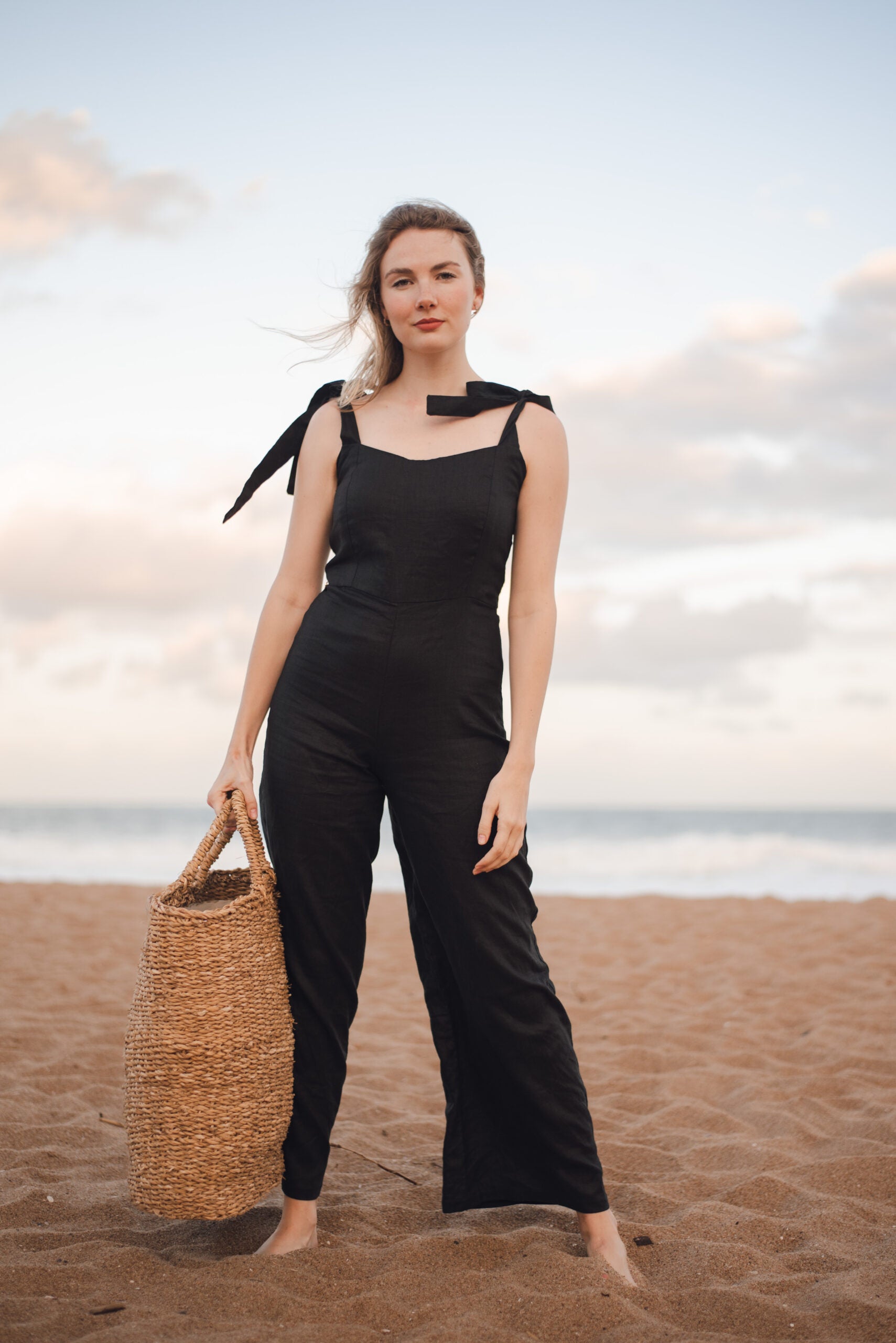 Elegant woman in black jumpsuit on beach, showcasing bespoke Linen clothing by South African designer.