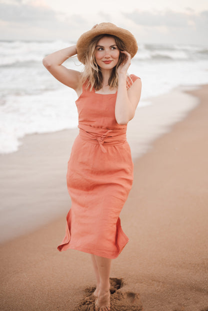 A stunning smiling woman in a vibrant peach dress and matching grass hat gracefully stands on the sandy beach, exuding elegance and style. Her attire, meticulously crafted by a renowned bespoke Linen clothing designer from South Africa, adds a touch of luxury to this picturesque scene.