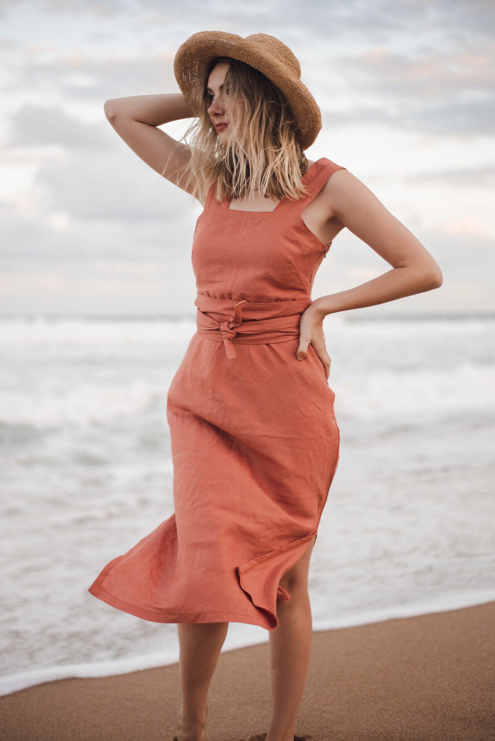 On the sun-kissed beach, a woman captivates with her allure, donning a striking peach dress and a chic hat. The impeccable attire, meticulously tailored by a distinguished Linen clothing designer and manufacturer based in South Africa, showcases the epitome of luxury and style.