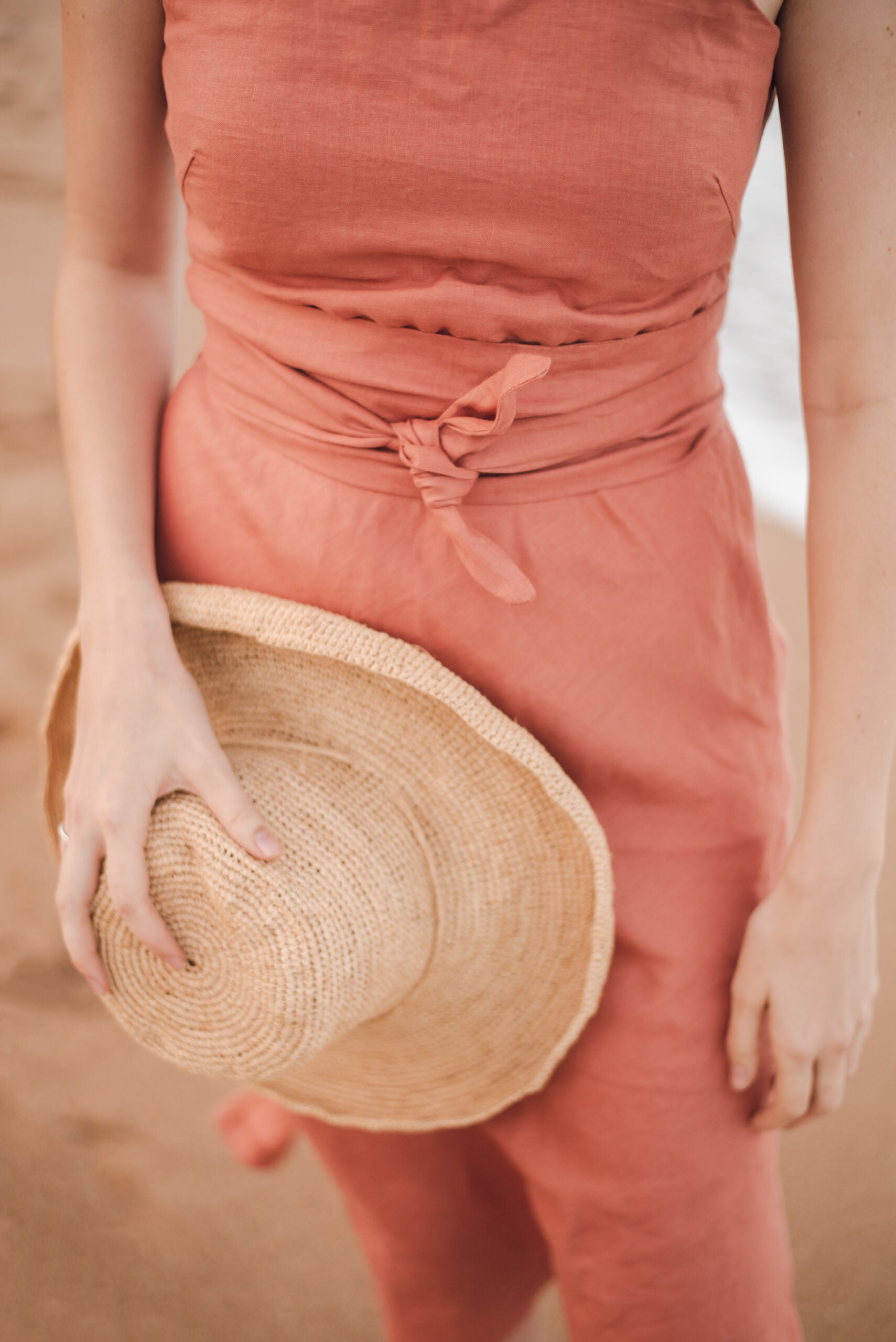 Hold her grass hat in hand, a stylish young woman wears a peach linen dress, as she stands with her feet on the ocean shoreline. Her clothing, meticulously tailored by a distinguished Linen clothing designer and manufacturer based in South Africa, showcases the height of luxury and style.