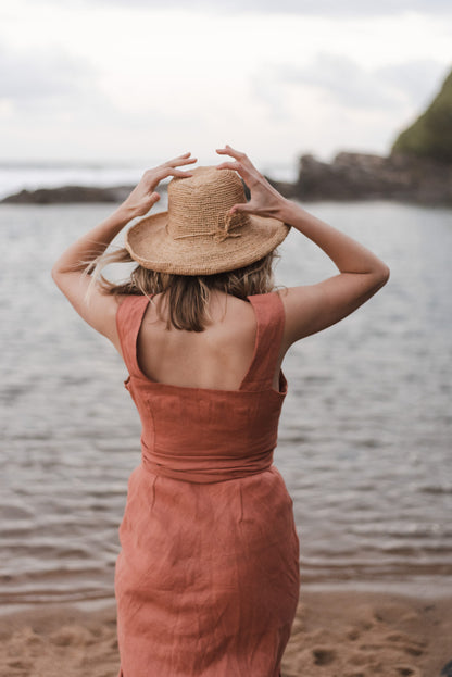 Adorned in a captivating peach dress and a fashionable hat, a woman photographed from behind radiates sophistication as she enjoys the serenity of the beach. The exquisite ensemble she wears is the creation of a prestigious Linen clothing designer and manufacturer hailing from South Africa, known for their bespoke craftsmanship.