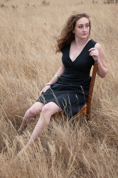 An elegant curly-haired woman in a sleek bespoke black linen dress seated on chair in South African field. The beautiful black dress handcrafted and designed in South Africa by renowned linen clothing designer.