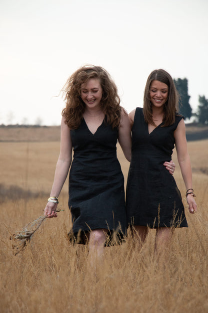 Two captivating woman, adorned in a sleek black dresses, gracefully walk amidst a picturesque field while hold one another close. Their exquisite attire by a renowned South African bespoke Linen clothing designer.