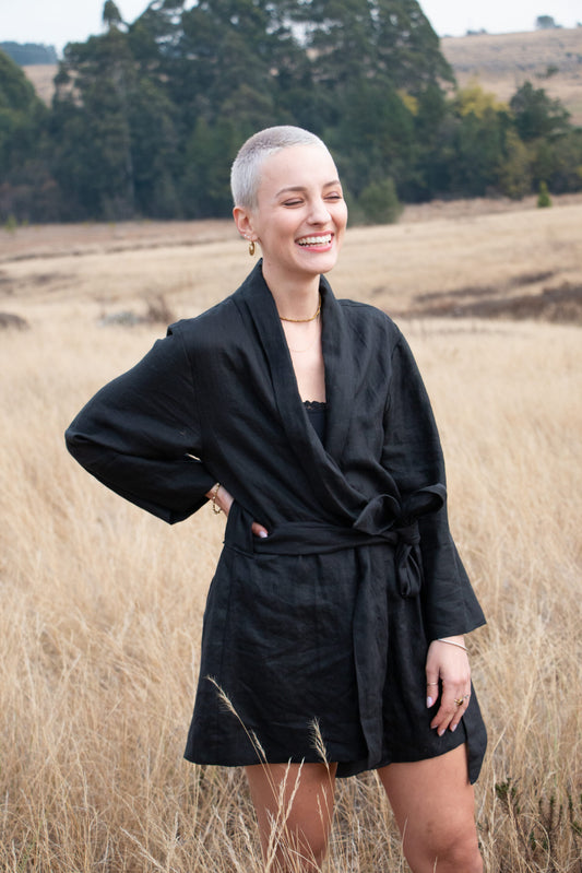 A stylish lady wearing a black linen kimono stands in a field of long grass. Her hand is on her waist as she smiles with her eyes closed. Her outfit is made from the highest quality linen fabric and represents the finest South African design and artistry.