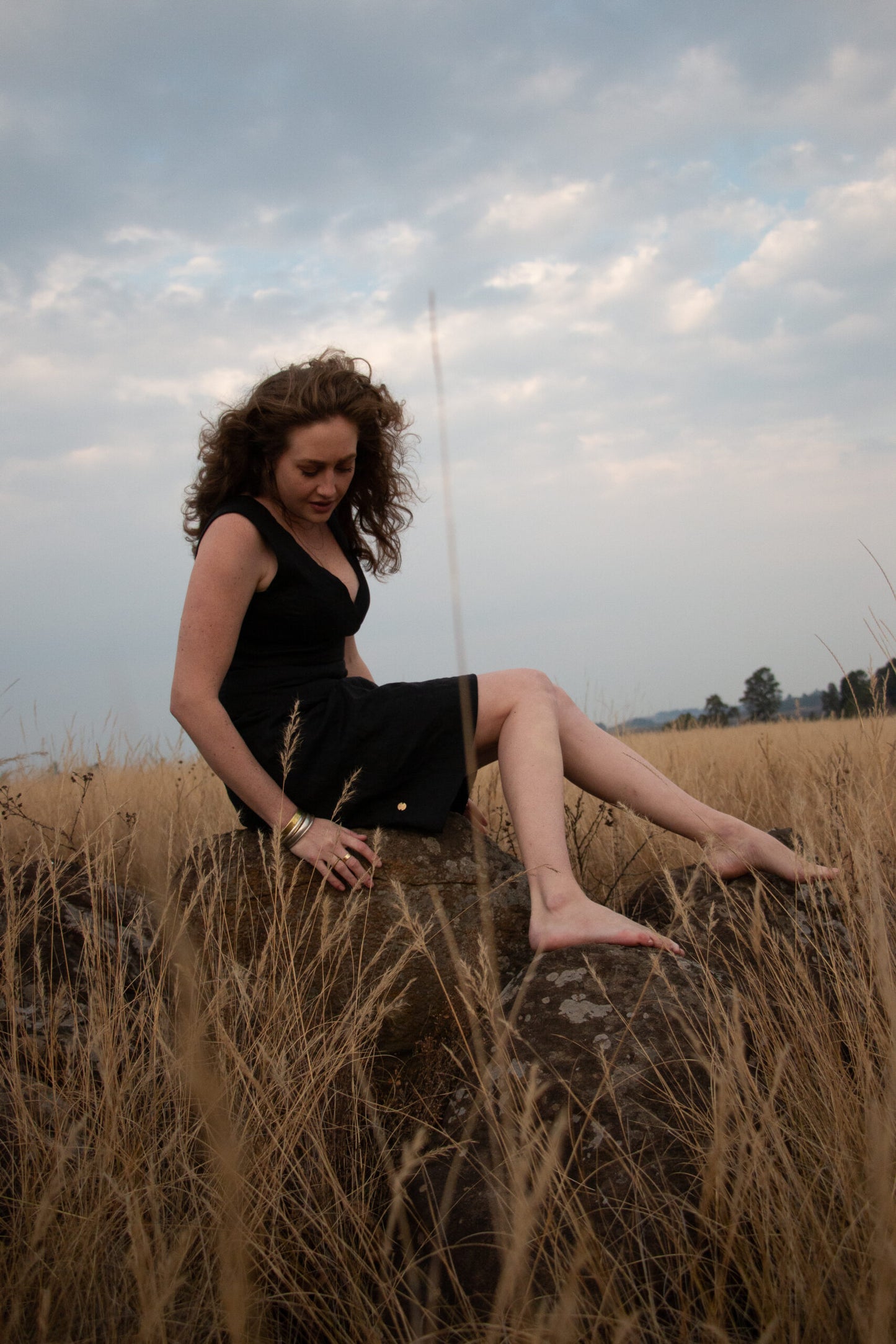 Sophisticated curl-haired woman in designer black linen dress, relaxing on rock in South African field. Her clothing designed and manufactured in South Africa, by bespoke linen clothing designer.