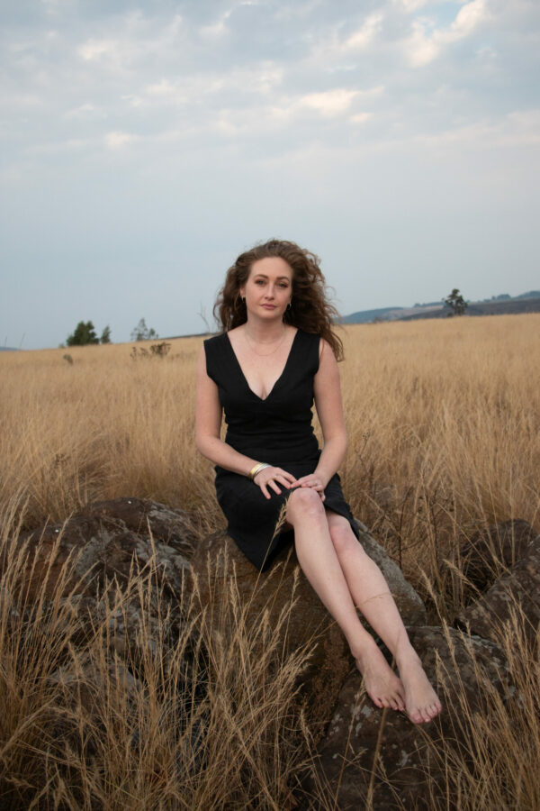 Elegant curly-haired woman in bespoke black linen dress seated on rock in South African field.
