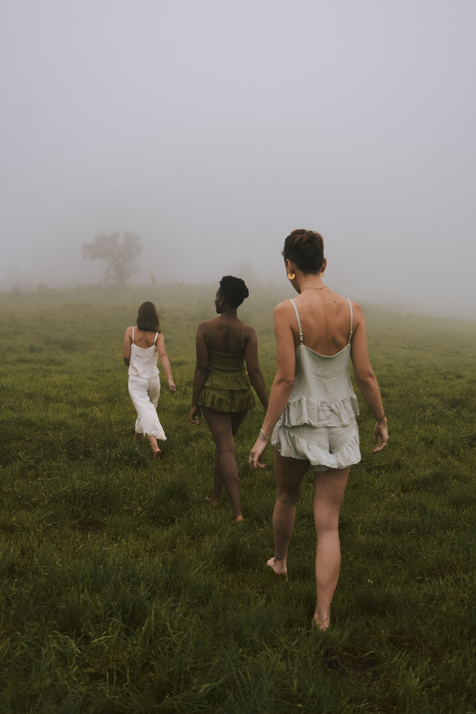 3 Women walk through a green field in single file on a misty day, all wearing lounge wear sets of clothing made out of luxury linen fabric.