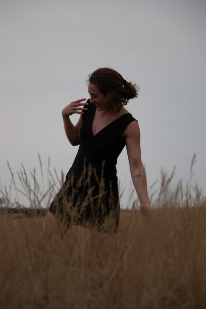 A Stylish woman wearing black dress walks in a field of tall grass. Her outfit is a bespoke Linen dress from a clothing designer and manufacturer in South Africa.