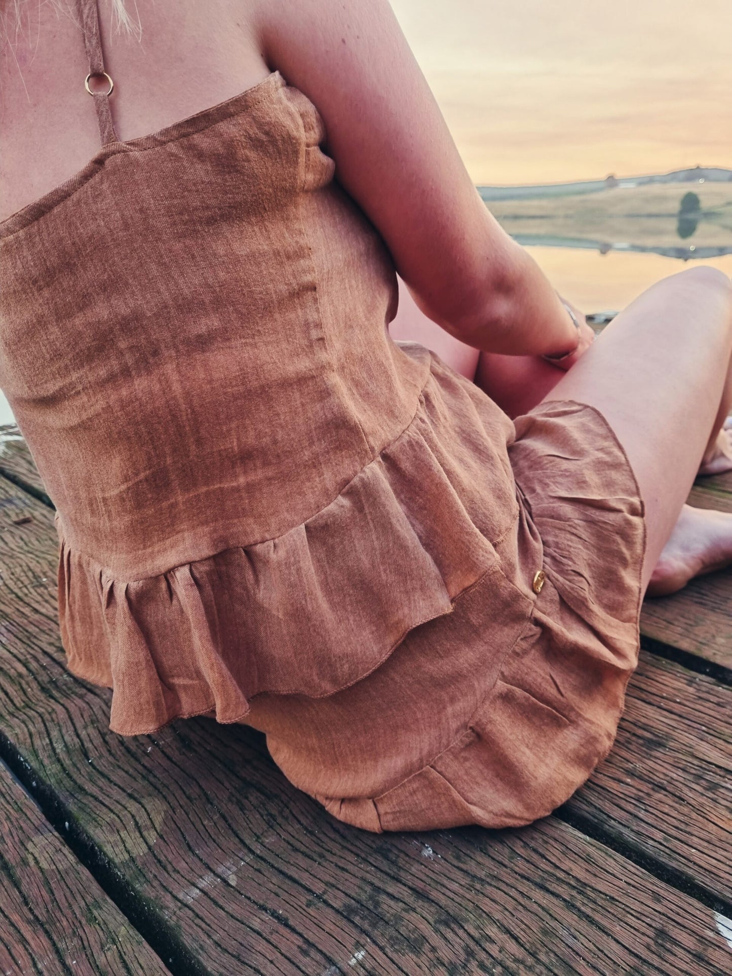 Woman sits on wooden deck wearing a rust coloured linen loungewear set that is made up of a linen top and linen short bottoms. Handcrafted from the highest quality linen fabric, the loungewear attire is designed and manufactured by hand in South Africa.