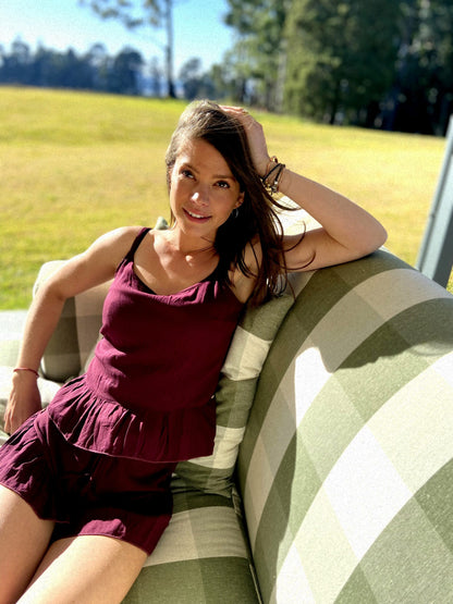 Woman relaxes in the sun on an outdoor couch while wearing a bespoke handcrafted linen loungewear set made up of a plum coloured linen top and plum coloured linen shorts. 