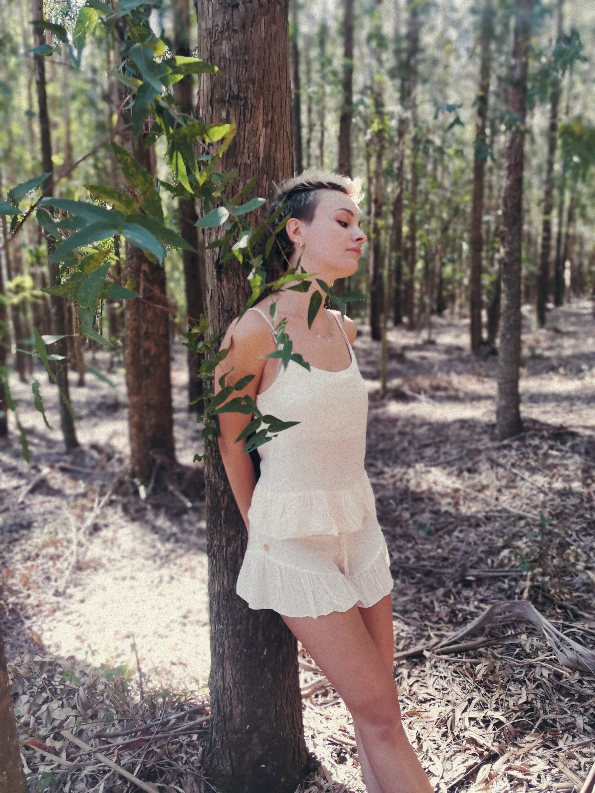 Classy woman leans against a tree trunk as she stands outdoors wearing a luxurious white linen loungewear top and bottom set. 