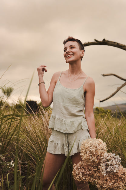 Beautiful woman stands holding flowers and wearing a sage green loungewear set of linen top and shorts.