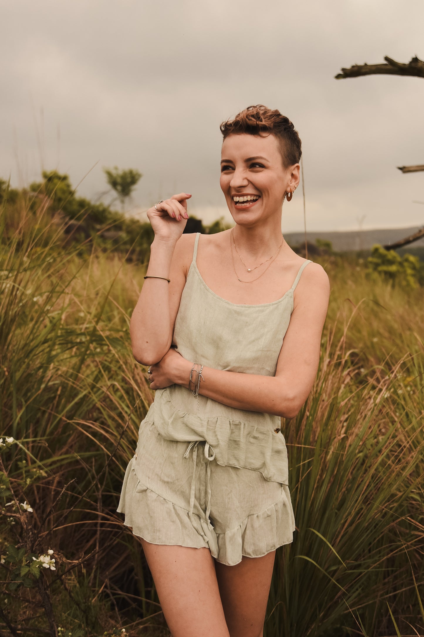 Smiling woman stand among tall grass wearing a comfortable linen loungewear set crafted out of sage green high quality pure linen fabric.