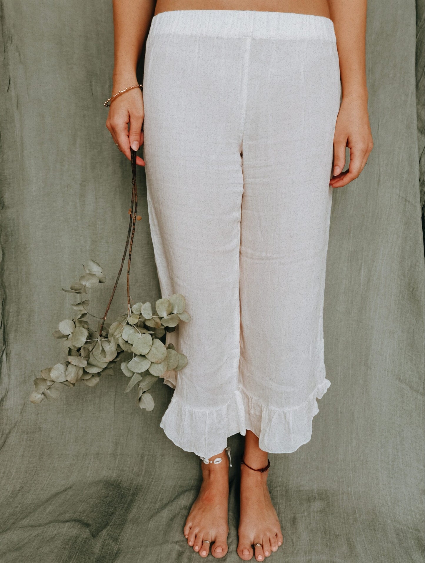 Dressed in comfortable white linen long pants, a woman stands in front of a sage background hold sage coloured foliage. Her pants are handcrafted from the finest linen fabric and are designed and manufactured in South Africa.