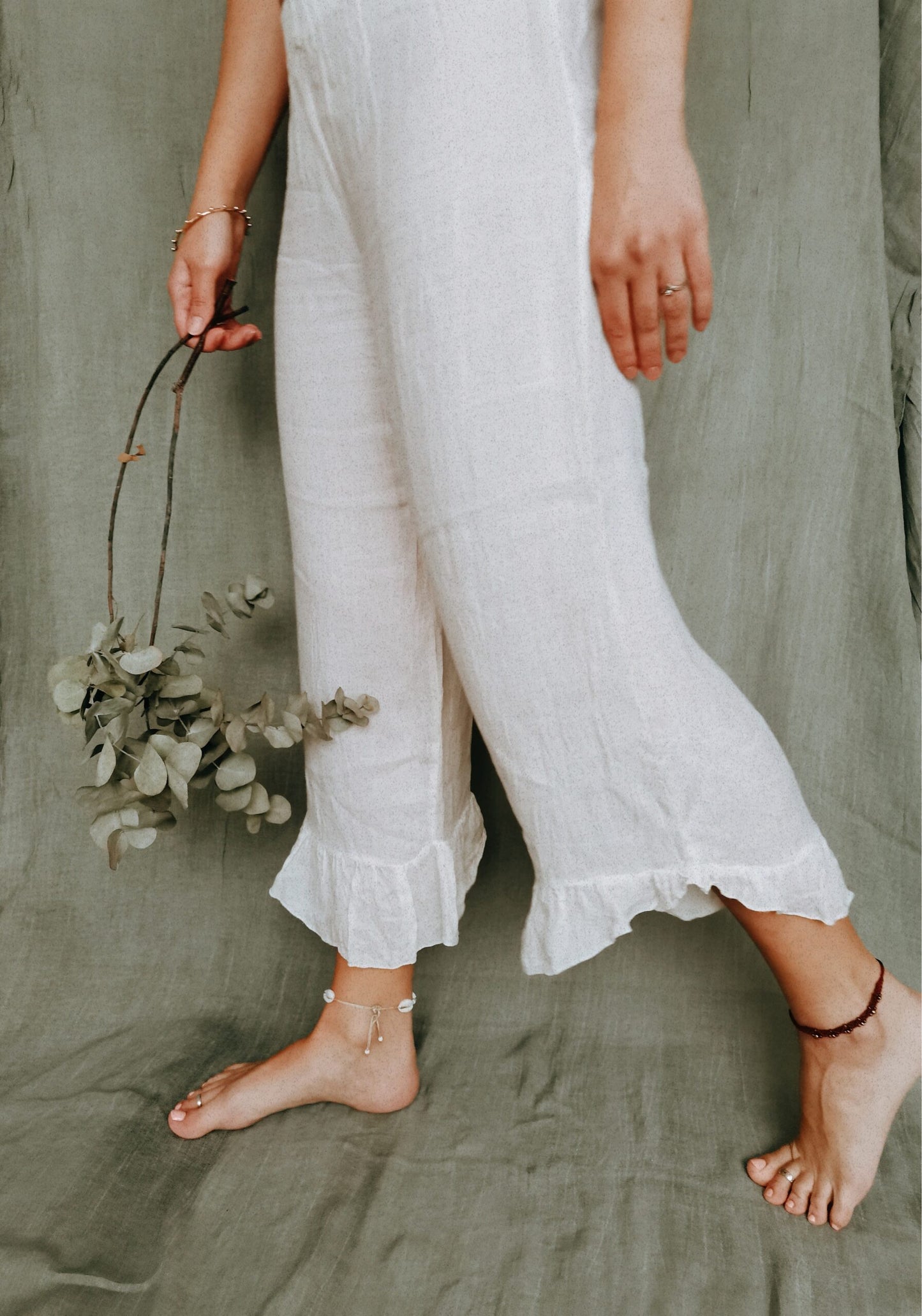 Standing elegantly, a woman wears light extra comfortable white linen long pants, as she stands in front of a sage green background.  