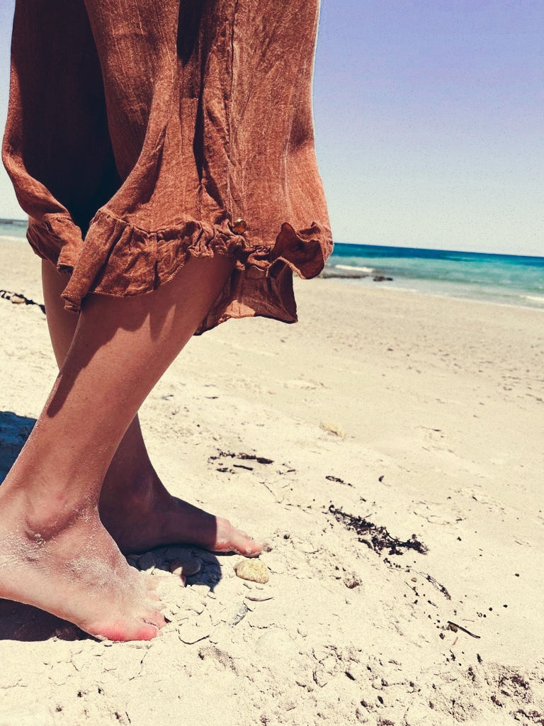 Standing on a sandy South African beach, a woman wears flowing rust coloured linen pants. Designed to be extra comfortable, her pants are crafted from the finest linen and designed and manufactured in South Africa