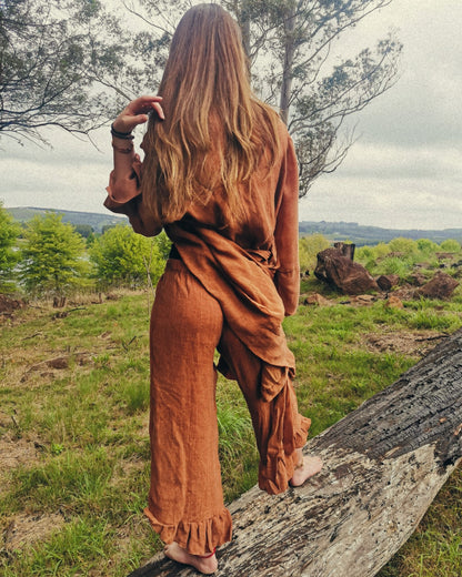 A woman stands outdoors ontop of a log wearing a rust coloured linen loungewear set made of up a linen top, linen long pants and a linen kimono.  Her outfit is designed and handmade by South African linen clothing designers Hare & Manuela.
