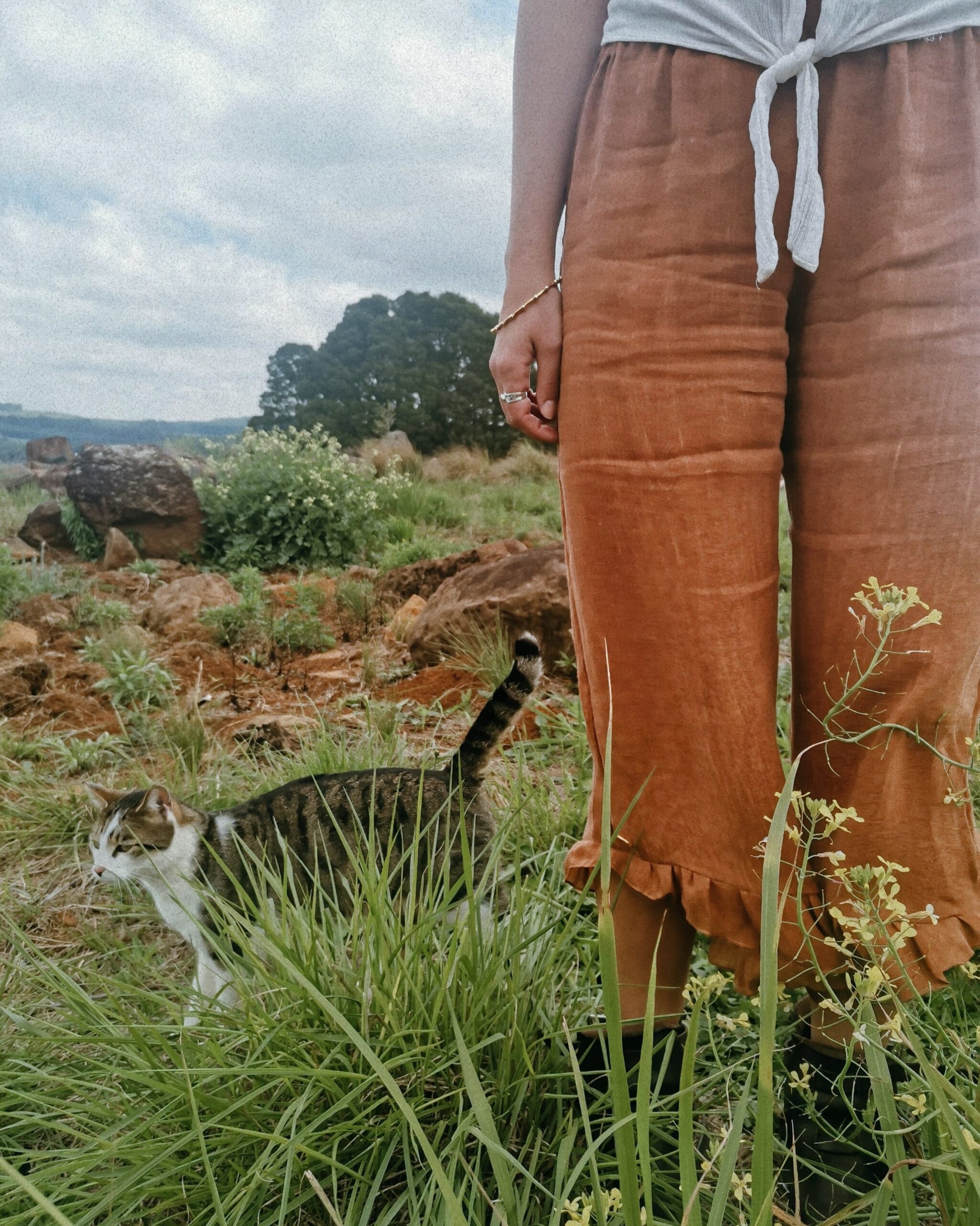 Standing in a field of green, a woman wears comfortable rust coloured linen fabric long pants. Design to be extra comfortable and made with the finest linen material, her long pants are designed and handmade in South Africa.